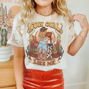Puede incluir: Una camiseta de color crema con un estampado gr&aacute;fico de dos figuras con sombreros de vaquero en un paisaje des&eacute;rtico con cactus y rosas. El texto "COW GIRL" aparece en la parte superior y "LIKE ME" en una cinta en la parte inferior.