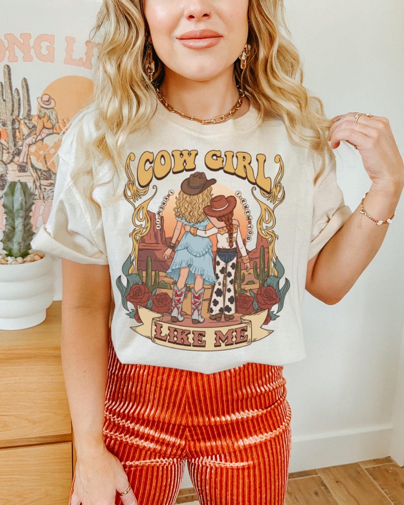 Puede incluir: Una camiseta de color crema con un estampado gr&aacute;fico de dos figuras con sombreros de vaquero en un paisaje des&eacute;rtico con cactus y rosas. El texto "COW GIRL" aparece en la parte superior y "LIKE ME" en una cinta en la parte inferior.