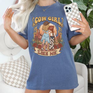 Puede incluir: Una camiseta azul de manga corta con un estampado gr&aacute;fico de dos figuras con sombreros de vaquero y ropa del oeste en un paisaje des&eacute;rtico con cactus y rosas. El texto COW GIRL LIKE ME est&aacute; impreso en una fuente decorativa.