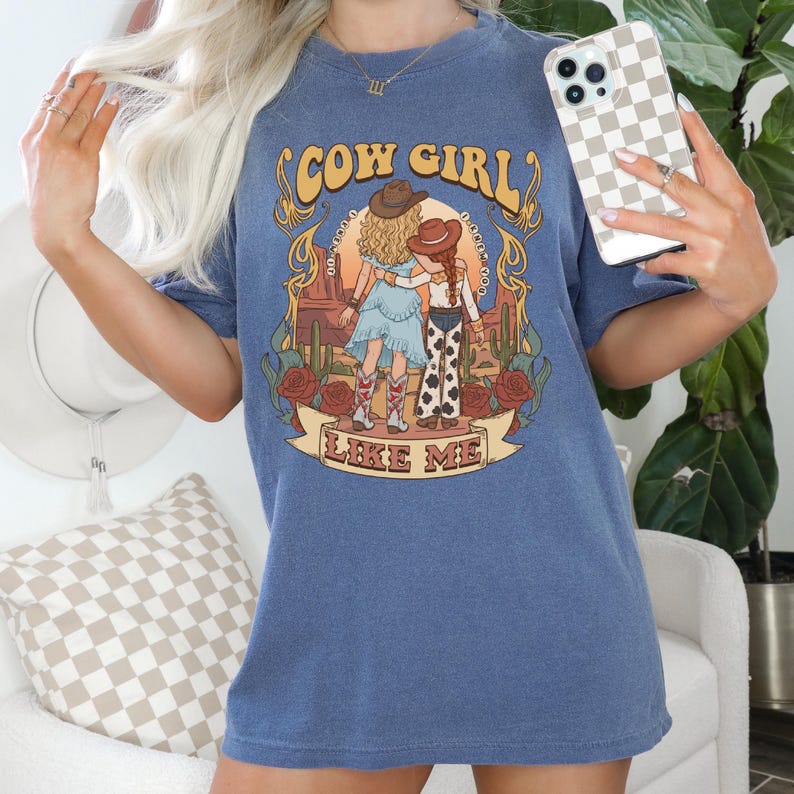 Puede incluir: Una camiseta azul de manga corta con un estampado gr&aacute;fico de dos figuras con sombreros de vaquero y ropa del oeste en un paisaje des&eacute;rtico con cactus y rosas. El texto COW GIRL LIKE ME est&aacute; impreso en una fuente decorativa.