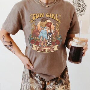 Puede incluir: Una camiseta marr&oacute;n de manga corta con un estampado gr&aacute;fico de dos figuras con vestimenta western en un paisaje des&eacute;rtico con cactus y rosas. El texto "COW GIRL" y "LIKE ME" est&aacute; impreso en una fuente retro sobre y bajo la ilustraci&oacute;n.