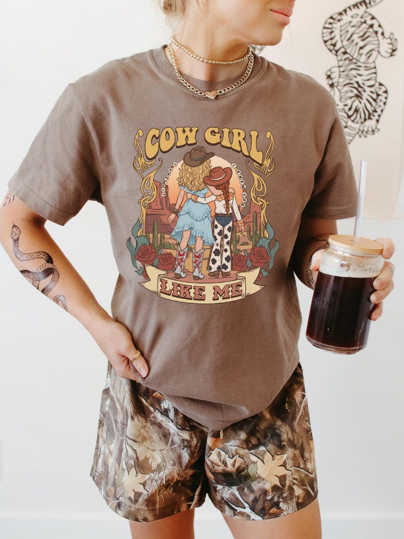 Puede incluir: Una camiseta marr&oacute;n de manga corta con un estampado gr&aacute;fico de dos figuras con vestimenta western en un paisaje des&eacute;rtico con cactus y rosas. El texto "COW GIRL" y "LIKE ME" est&aacute; impreso en una fuente retro sobre y bajo la ilustraci&oacute;n.