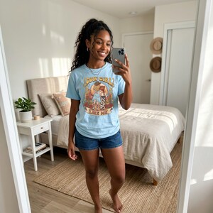 Puede incluir: Una camiseta de manga corta de color azul claro con un estampado gr&aacute;fico de dos figuras con sombreros y botas de vaquero, junto al texto "COW GIRL LIKE ME" en una fuente estilizada, combinada con unos pantalones cortos de mezclilla azul.