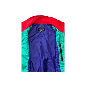 K&ouml;nnte beinhalten: Eine blaue, gr&uuml;ne und rote Jacke mit einer Rei&szlig;verschlusstasche und einem schwarzen Etikett mit der Aufschrift "YAMAHA" auf der Innenseite.