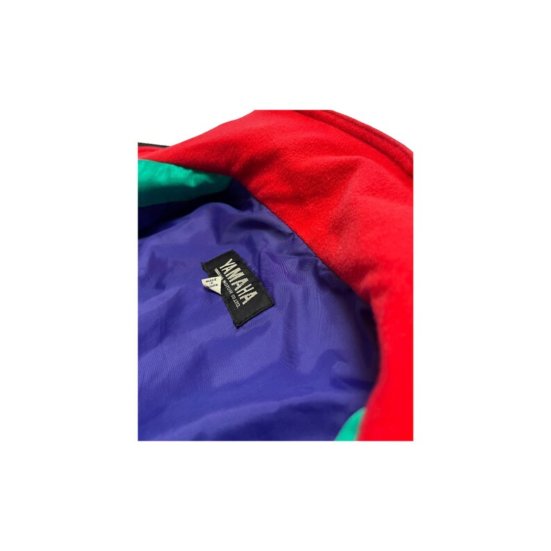 K&ouml;nnte beinhalten: Nahaufnahme einer blauen, roten und gr&uuml;nen Jacke mit einem Yamaha-Logo-Etikett. Das Etikett lautet "Yamaha Motor Co. Ltd."
