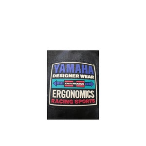 K&ouml;nnte beinhalten: Ein schwarzer Patch mit wei&szlig;em, blauem und rotem Text, der "Yamaha Designer Wear Ergonomics Racing Sports" lautet. Der Patch hat einen blauen und wei&szlig;en Pfeil, der nach rechts zeigt, und einen blauen und wei&szlig;en Pfeil, der nach links zeigt.