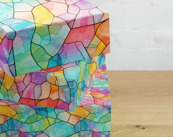 May include: Three stacked gift boxes with a colourful stained glass pattern. The boxes feature a vibrant design with shades of pink, yellow, blue, and green, outlined in black. The boxes are on a wooden surface.