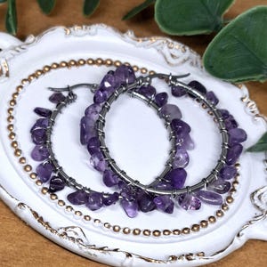 Könnte beinhalten: Ein Paar Creolen mit Amethyststeinen, die um die Creolen gewickelt sind. Die Ohrringe sind silberfarben und die Amethyststeine sind tiefviolett.