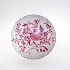 May include: A white ceramic lid with a pink floral design. The lid is round and has a raised edge. The floral design is intricate and features a variety of flowers and leaves.