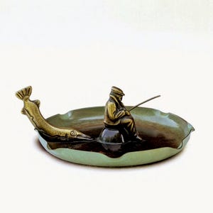 May include: An ornamental ashtray depicting a fisherman in a boat, with a fish-shaped detail. The ashtray has a green and brown colour scheme, with a bronze-coloured fisherman and fish. The fisherman is holding a fishing rod.
