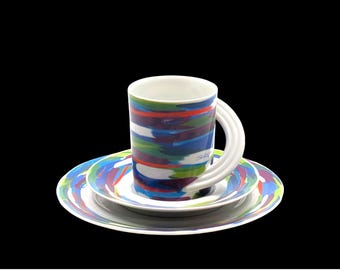 May include: A white ceramic coffee cup with a colourful abstract design in shades of blue, green, red, and purple. The cup has a unique double handle and is sitting on a matching saucer.