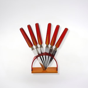 May include: A set of six red and orange butter knives with silver blades, arranged in a fan shape in a wooden stand.