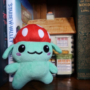 May include: A plush toy shaped like a mushroom with a green body, red cap with white spots, and a smiling face. The toy is sitting on a shelf with books and a ceramic house.