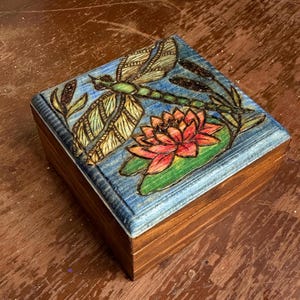May include: A wooden box with a detailed, hand-painted design. The lid features a dragonfly, a lotus flower, and aquatic plants against a blue background. The box is square and has a natural wood finish.