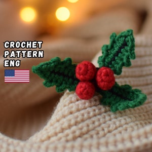May include: A crocheted holly sprig with green leaves and red berries. The text "CROCHET PATTERN ENG" is visible. The holly is placed on a beige knitted fabric. The background has blurred warm-toned lights.