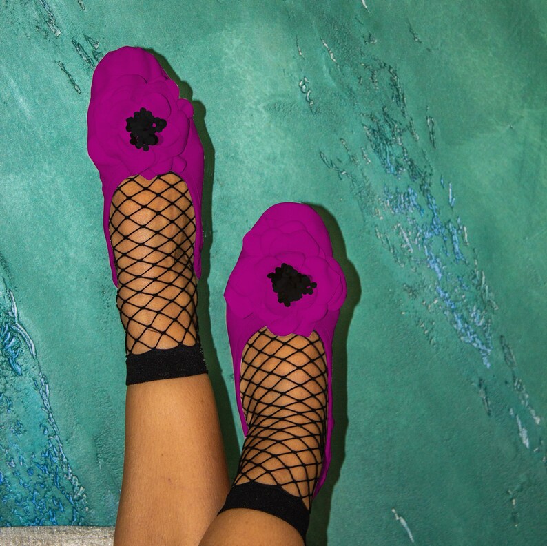 May include: A pair of pink ballet flats with black floral embellishments and fishnet stockings. The flats are on a person's feet.