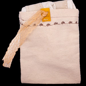 May include: A beige canvas bag with a brown embroidered border and a small yellow leather tag. The bag is closed and has a lace trim hanging from the top.