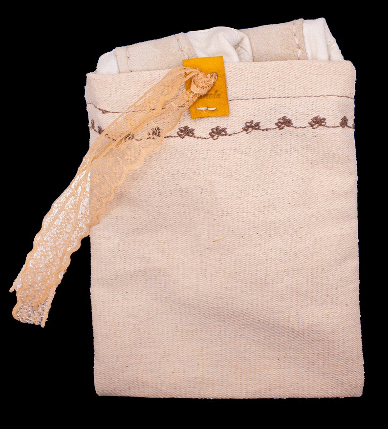 May include: A beige canvas bag with a brown embroidered border and a small yellow leather tag. The bag is closed and has a lace trim hanging from the top.
