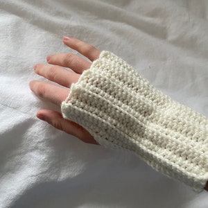 Puede incluir: Guantes de ganchillo sin dedos de color crema. Los guantes están hechos con un punto texturizado, y la parte de la muñeca es ligeramente más ancha que la abertura de la mano. Los guantes se muestran en una mano.