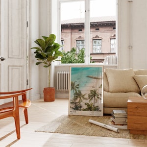 May include: A framed beach scene print with palm trees and ocean waves leans against a beige sofa in a bright room. A large potted plant and a wooden chair are also visible. The print has the word "BEACH" at the bottom.