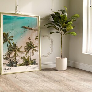 May include: A framed beach print with a gold-colored frame. The image shows a tropical beach scene with palm trees, turquoise water, and people. The word "BEACH" is printed at the bottom of the frame.
