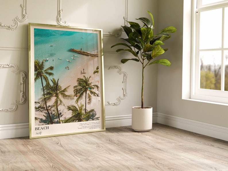 May include: A framed beach print with a gold-colored frame. The image shows a tropical beach scene with palm trees, turquoise water, and people. The word "BEACH" is printed at the bottom of the frame.