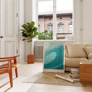 May include: A framed poster with a photograph of a large wave crashing. The poster is hanging on a white wall in a living room with a beige couch, a wooden coffee table, and a potted plant.