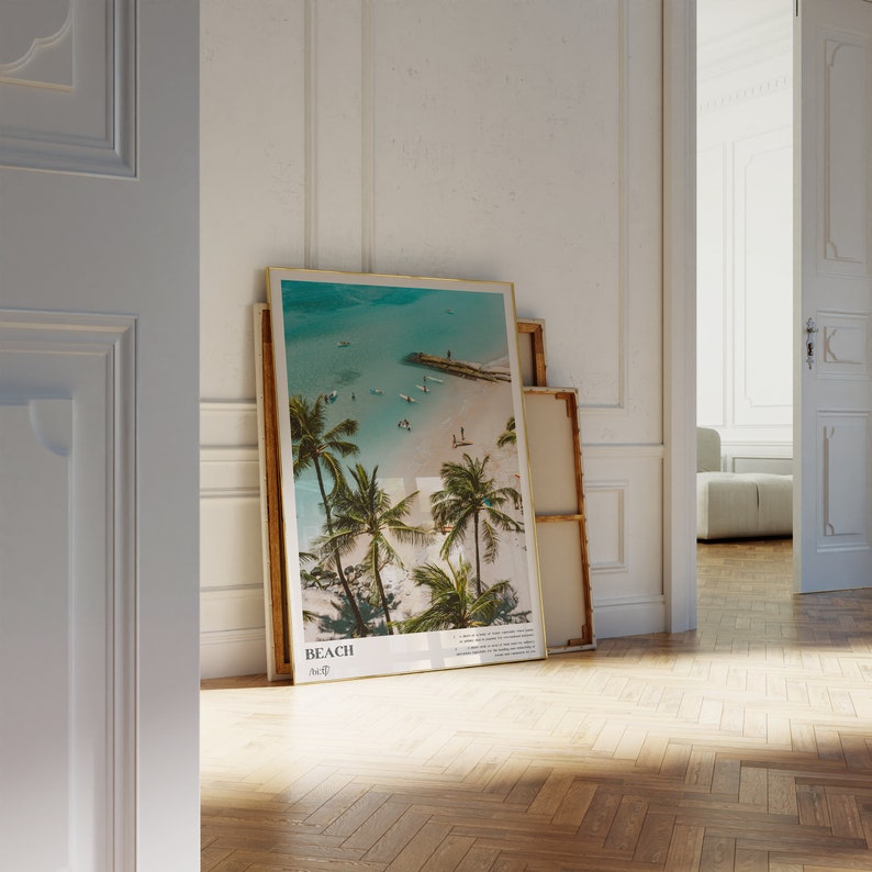 May include: A framed beach scene print featuring a turquoise ocean, sandy shore, and palm trees. The print has a gold frame and the word "BEACH" is printed at the bottom. Other prints are leaning against the wall.