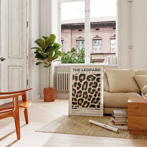 May include: A framed poster with a leopard print design. The poster is titled "The Leopard Wild & Sexy". The poster is hanging on a white wall in a living room with a brown rug, a wooden chair, a couch, and a plant.