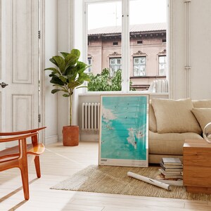 May include: A framed poster with a blue and white image of surfers riding waves. The poster is hanging on a white wall in a living room with a beige couch, a wooden coffee table, and a potted plant.