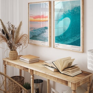 May include: Two framed prints of ocean waves, one with a sunset and the other with a breaking wave. The prints are hanging on a white wall above a wooden console table with books and a vase of dried flowers.