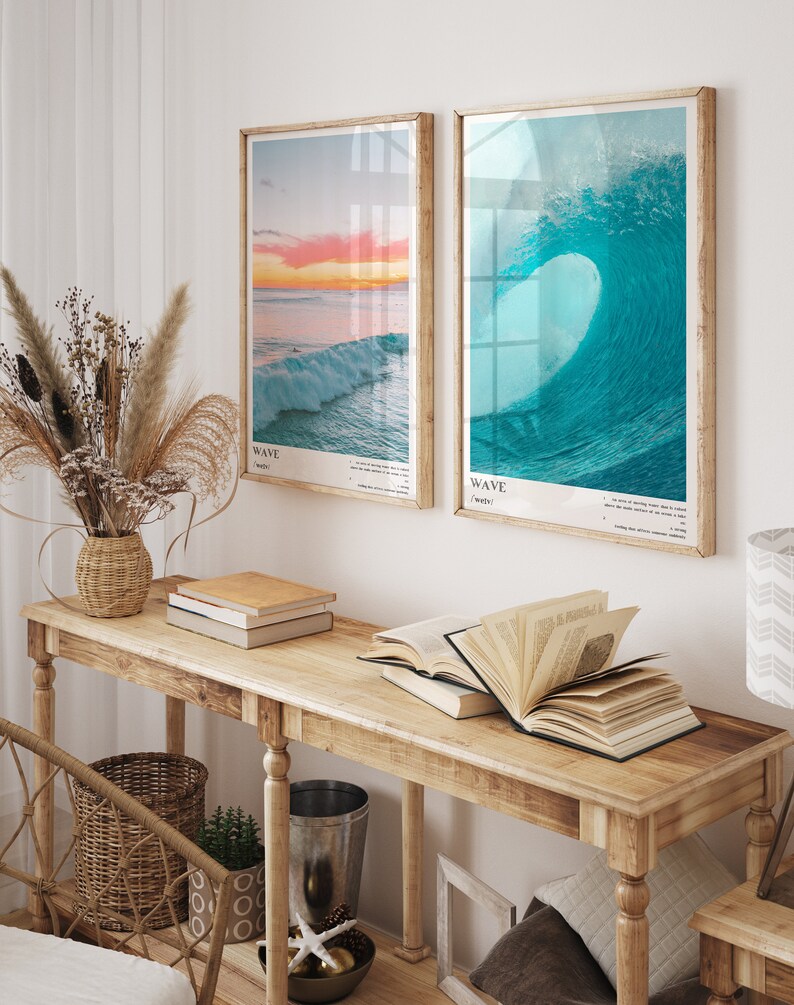 May include: Two framed prints of ocean waves, one with a sunset and the other with a breaking wave. The prints are hanging on a white wall above a wooden console table with books and a vase of dried flowers.