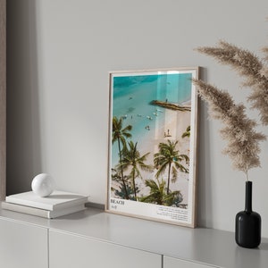 May include: A framed beach print with a light wood frame. The image shows a tropical beach scene with palm trees, turquoise water, and people. The print has the word "BEACH" and other text at the bottom.