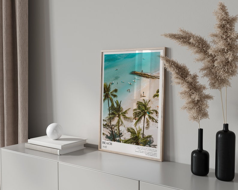 May include: A framed beach print with a light wood frame. The image shows a tropical beach scene with palm trees, turquoise water, and people. The print has the word "BEACH" and other text at the bottom.