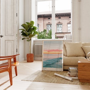 May include: A framed poster with a photograph of a wave crashing in the ocean at sunset. The poster is hanging on a white wall in a living room with a beige couch, a wooden coffee table, and a potted plant.