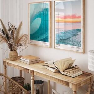 May include: Two framed prints of ocean waves, one with a blue wave and the text "WAVE / wave", and the other with a sunset over the ocean and the text "WAVE / waves". The prints are hanging on a white wall above a wooden console table with books and a vase of dried flowers.
