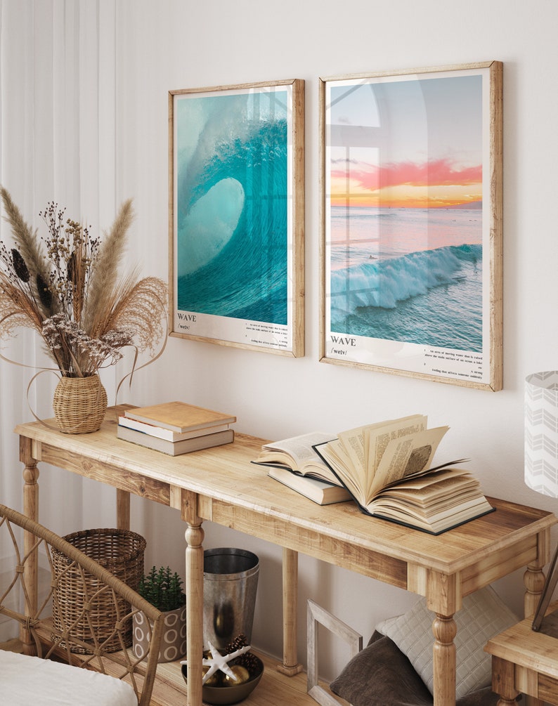May include: Two framed prints of ocean waves, one with a blue wave and the text "WAVE / wave", and the other with a sunset over the ocean and the text "WAVE / waves". The prints are hanging on a white wall above a wooden console table with books and a vase of dried flowers.