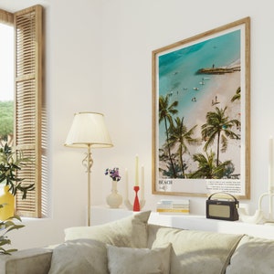 May include: A framed beach scene print with a wooden frame. The print features a turquoise ocean, white sand, and palm trees. The word "BEACH" is printed at the bottom. The print is displayed above a sofa with beige cushions.