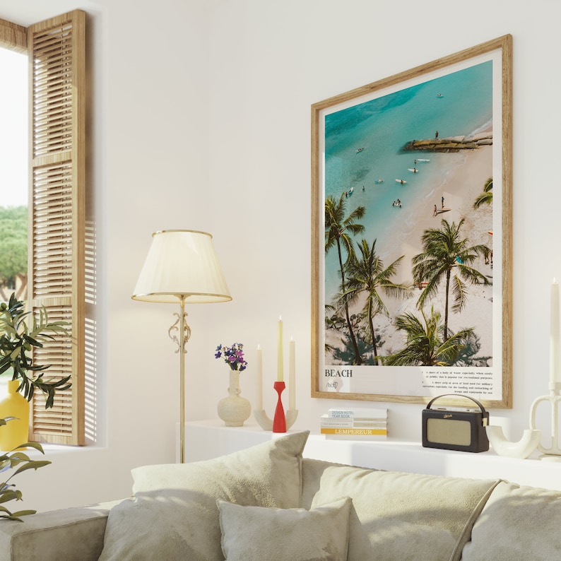 May include: A framed beach scene print with a wooden frame. The print features a turquoise ocean, white sand, and palm trees. The word "BEACH" is printed at the bottom. The print is displayed above a sofa with beige cushions.