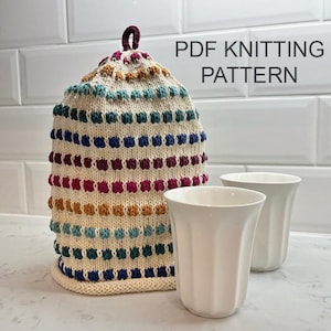 Puede incluir: Un calienta-té de punto de colores con un patrón a rayas y un lazo de hilo marrón en la parte superior. El calienta-té está sobre una encimera blanca junto a dos tazas de cerámica blanca. El texto "PDF KNITTING PATTERN" está escrito en negro en la parte superior de la imagen.
