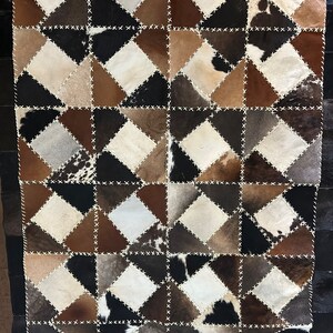 May include: A rectangular rug with a patchwork design. The rug features diamond-shaped patches in various shades of brown, black, and cream, stitched together with white thread. The rug's border is a solid black.
