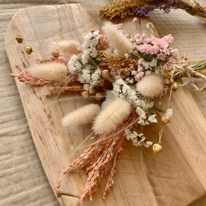 Puede incluir: Un ramo de flores secas en tonos blancos, rosas y beige. El ramo incluye colas de conejo blancas y esponjosas, peque&ntilde;as flores blancas y flores rosas. El ramo est&aacute; dispuesto sobre una superficie de madera.