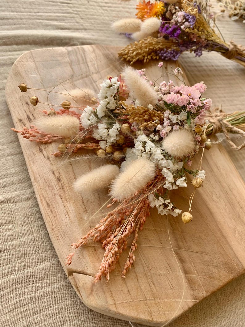 Puede incluir: Un ramo de flores secas en tonos blancos, rosas y beige. El ramo incluye colas de conejo blancas y esponjosas, peque&ntilde;as flores blancas y flores rosas. El ramo est&aacute; dispuesto sobre una superficie de madera.