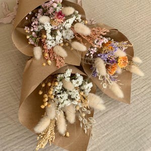 Puede incluir: Tres peque&ntilde;os ramos de flores secas envueltos en papel marr&oacute;n. Los ramos incluyen flores blancas, rosas, moradas y naranjas, as&iacute; como hierbas secas y vainas de semillas blancas y esponjosas.