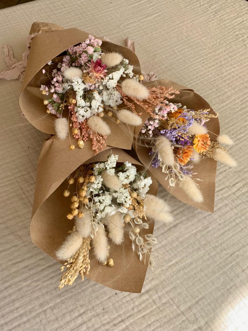 Puede incluir: Tres peque&ntilde;os ramos de flores secas envueltos en papel marr&oacute;n. Los ramos incluyen flores blancas, rosas, moradas y naranjas, as&iacute; como hierbas secas y vainas de semillas blancas y esponjosas.