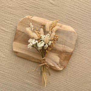 Puede incluir: Un peque&ntilde;o ramo de flores secas en tonos blancos y beige, atado con cuerda, sobre una superficie de madera.