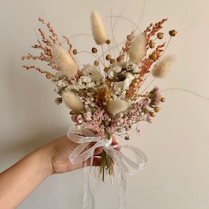 Puede incluir: Un ramo de flores secas en tonos blancos, rosas y beige. El ramo incluye hierbas secas, hierba de la pampa y aliento de beb&eacute;. Est&aacute; atado con una cinta blanca.