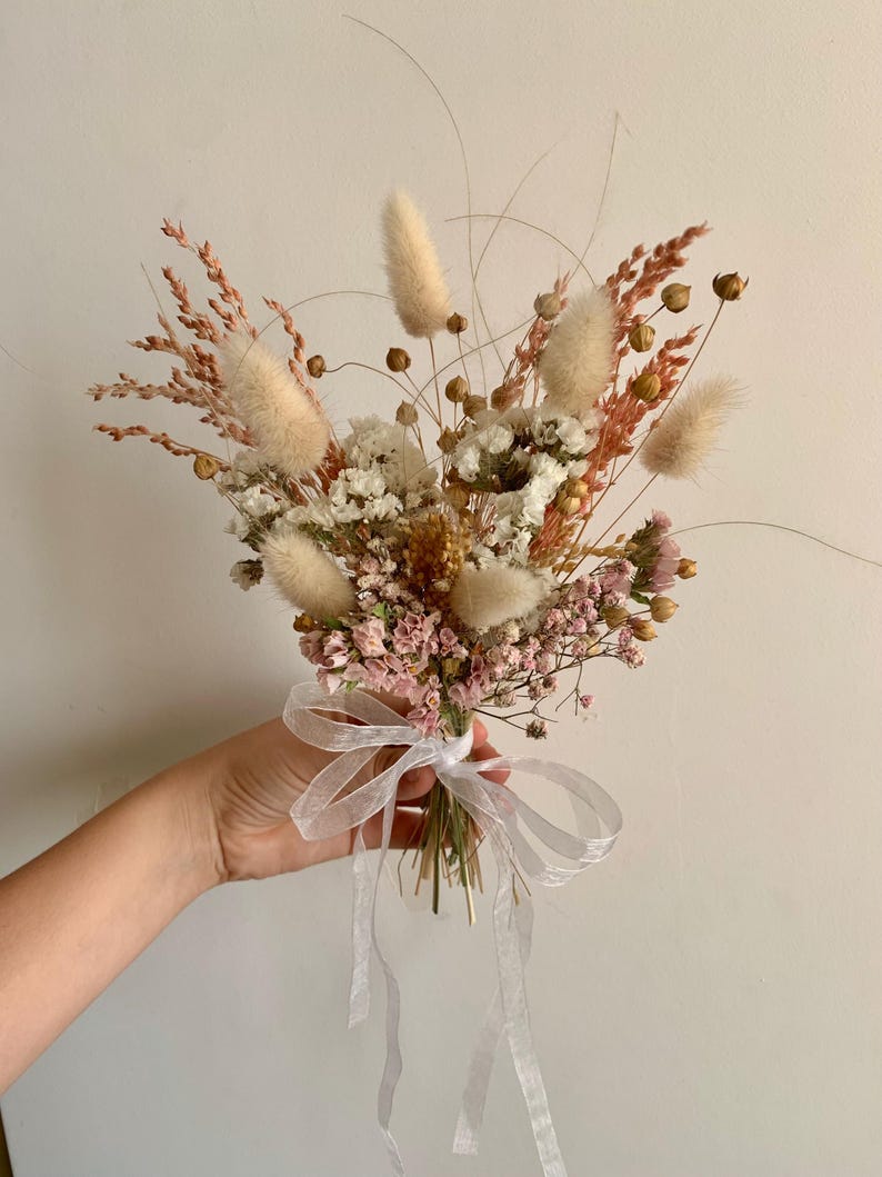 Puede incluir: Un ramo de flores secas en tonos blancos, rosas y beige. El ramo incluye hierbas secas, hierba de la pampa y aliento de beb&eacute;. Est&aacute; atado con una cinta blanca.