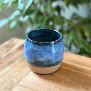 May include: A handcrafted ceramic tumbler with a rounded form. The upper section is glazed in varying shades of blue, blending into a natural tan base. The interior is a deep blue. The tumbler rests on a wooden surface.