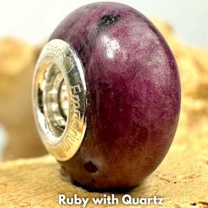 Könnte beinhalten: Eine runde, polierte Perle aus tief magenta-farbenem Rubin mit Quarzeinschlüssen. Die Perle verfügt über einen zentralen Kern aus silberfarbenem Metall mit dem eingravierten Text "Beads Emporium" am Rand und einer glatten, glänzenden Oberfläche.